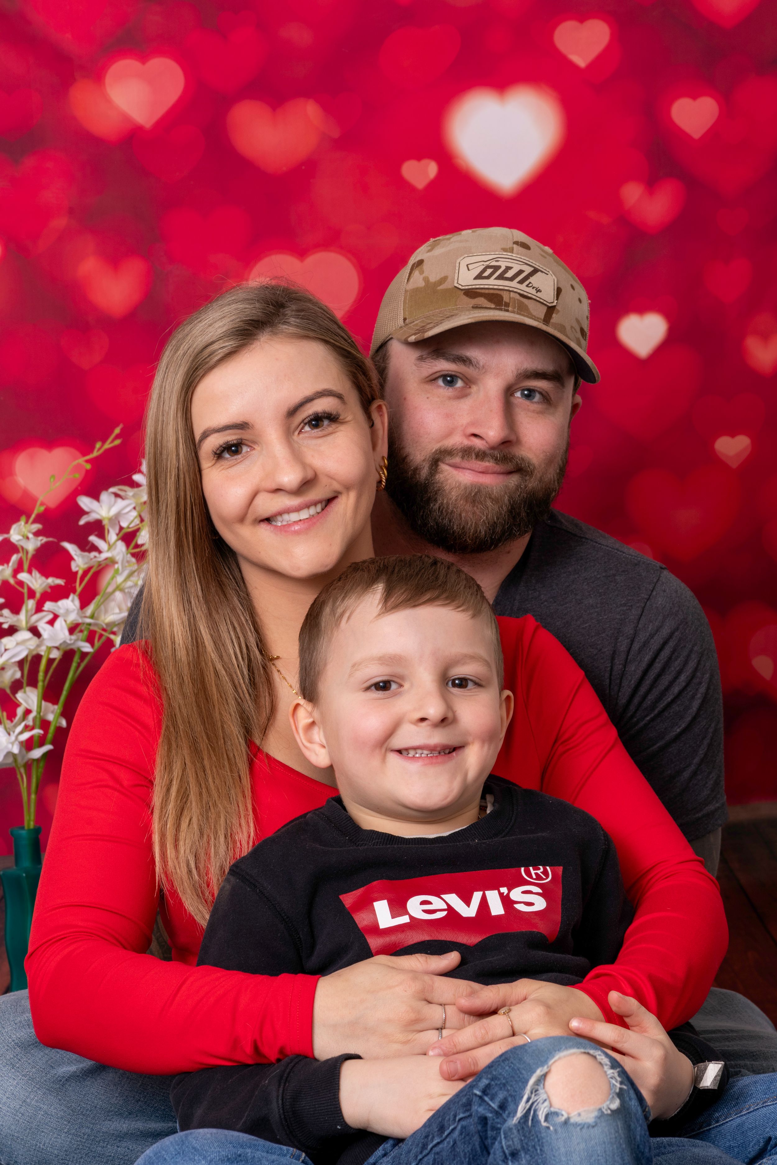 Family portrait with woman, bearded man in camo cap, and young boy in Levis shirt posed before red heart bokeh backdrop.