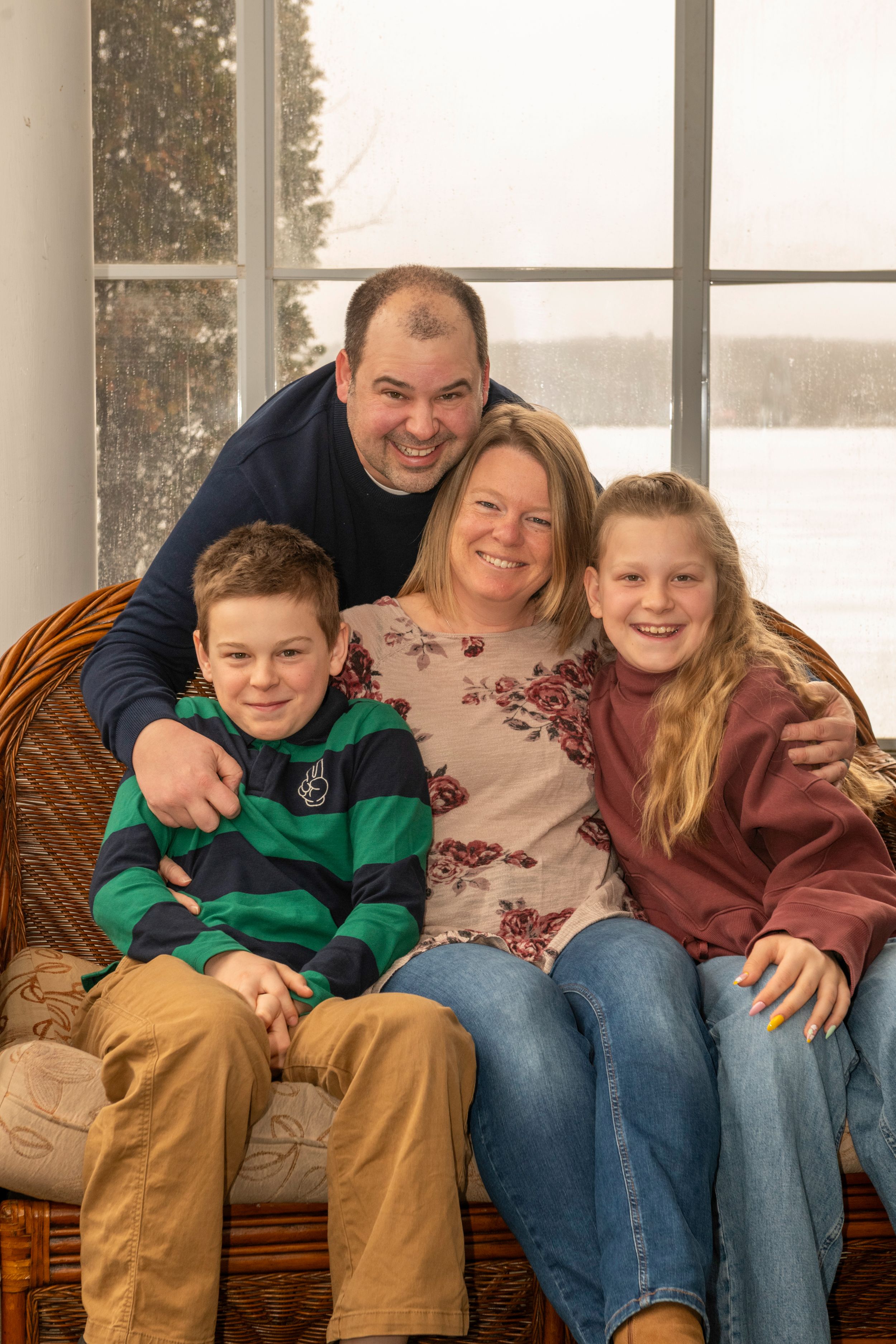 Happy family of four posing together on a wicker chair in front of a snowy winter window backdrop.