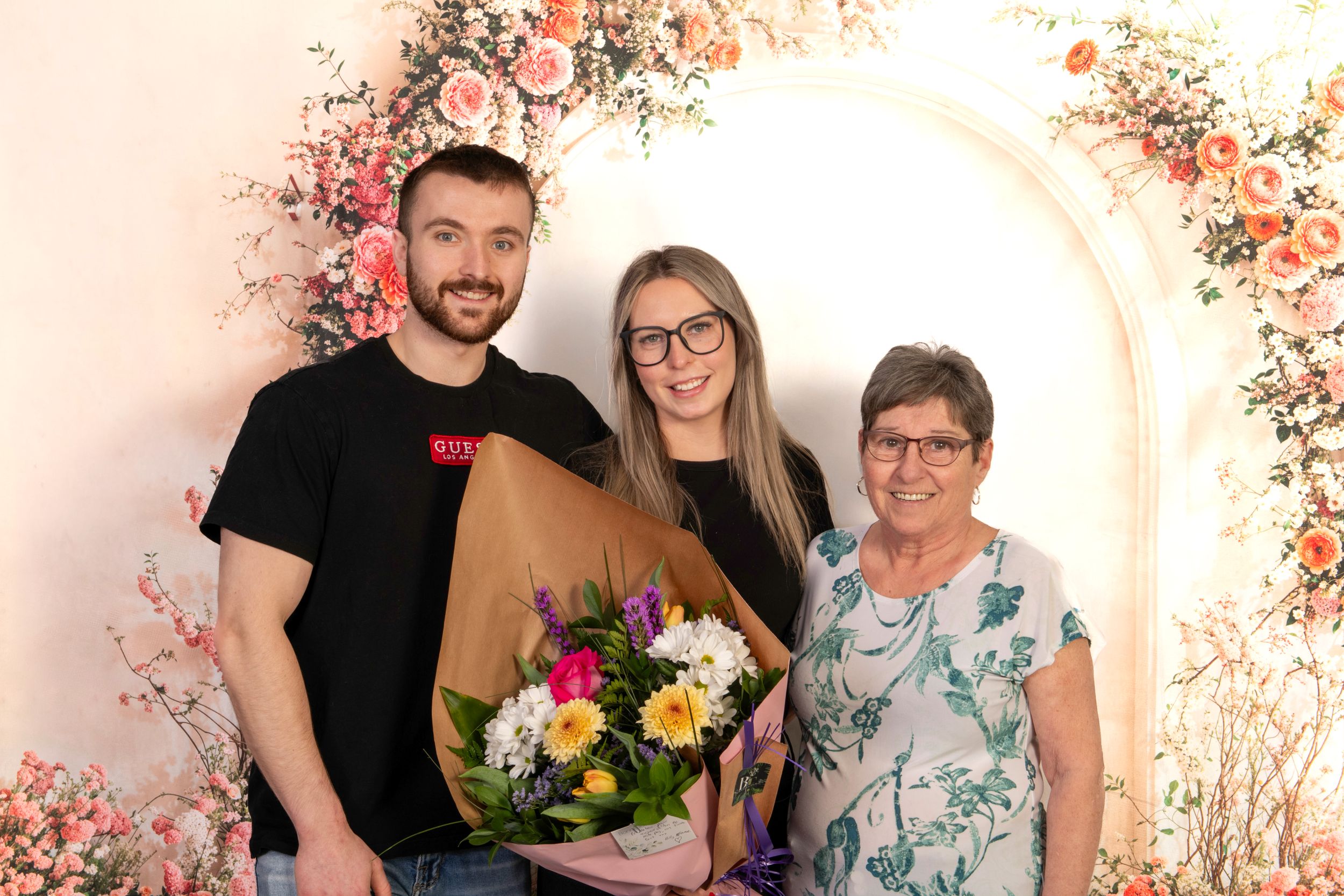 Three people posing together in front of a floral arch, with a woman holding a colorful bouquet of fresh flowers.