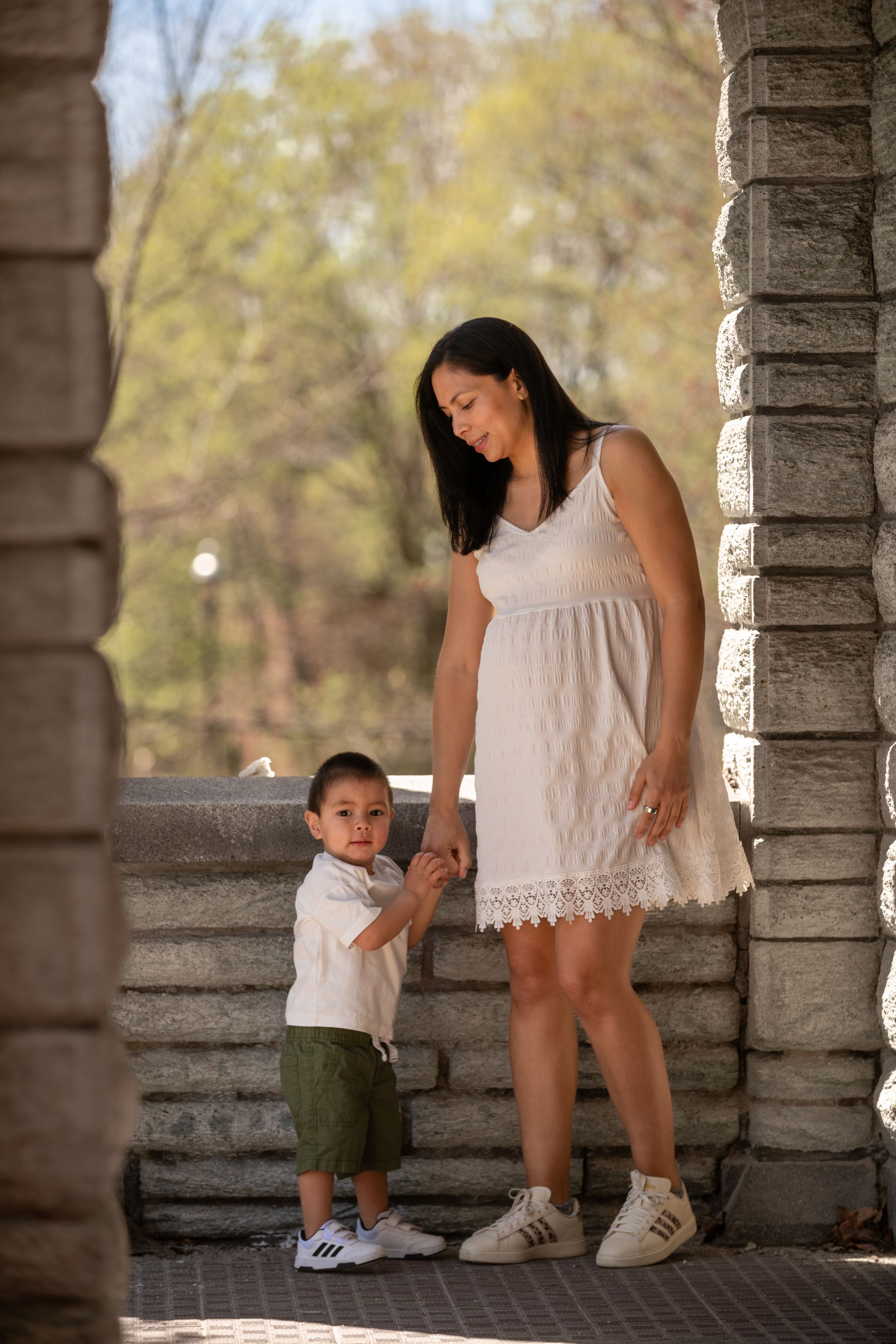 A mother in a white dress holds hands with her young son in a park near stone columns on a sunny day.