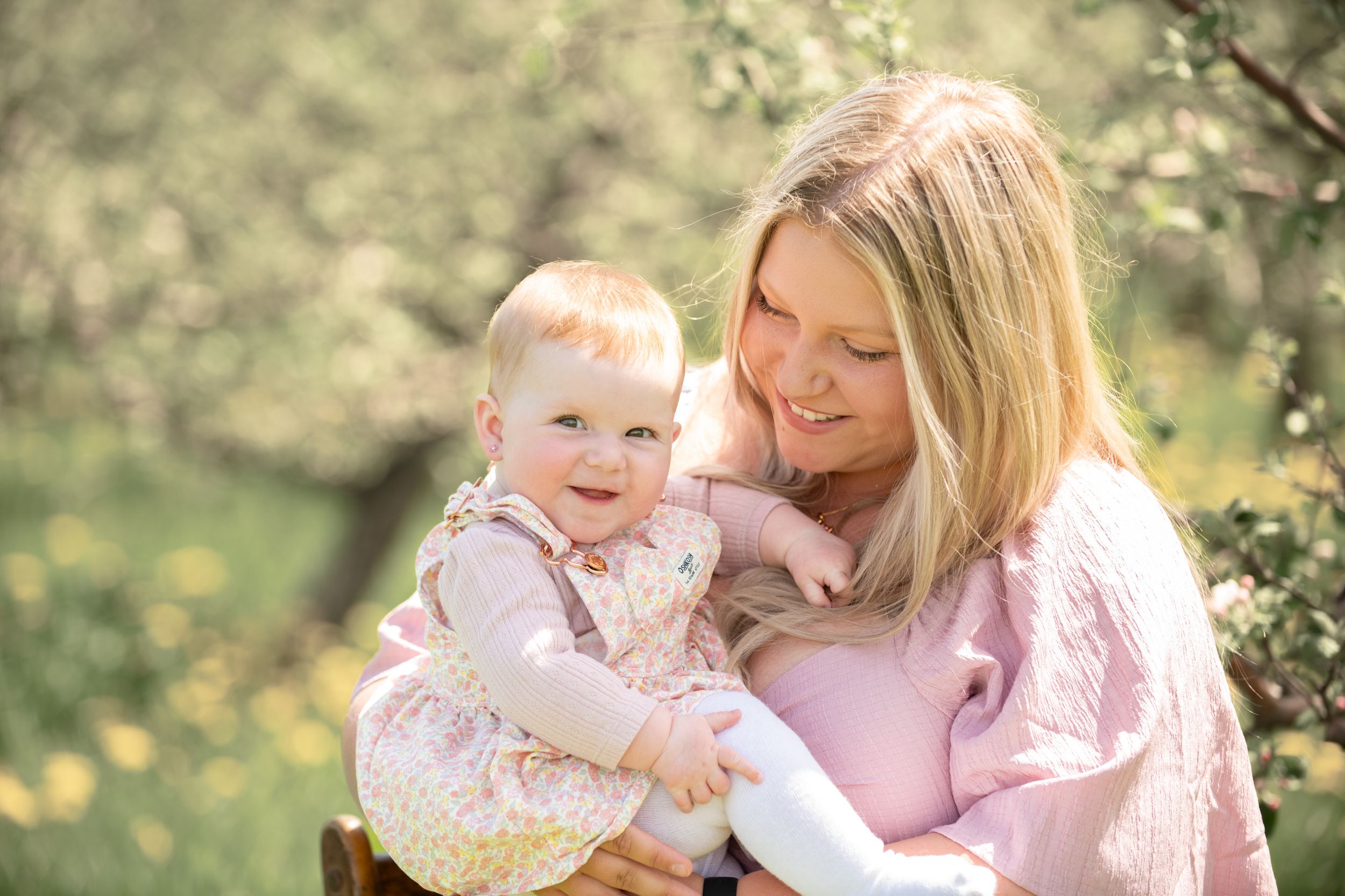 A smiling mother in pink holds her happy baby girl outdoors among blooming spring trees in soft sunlight.
