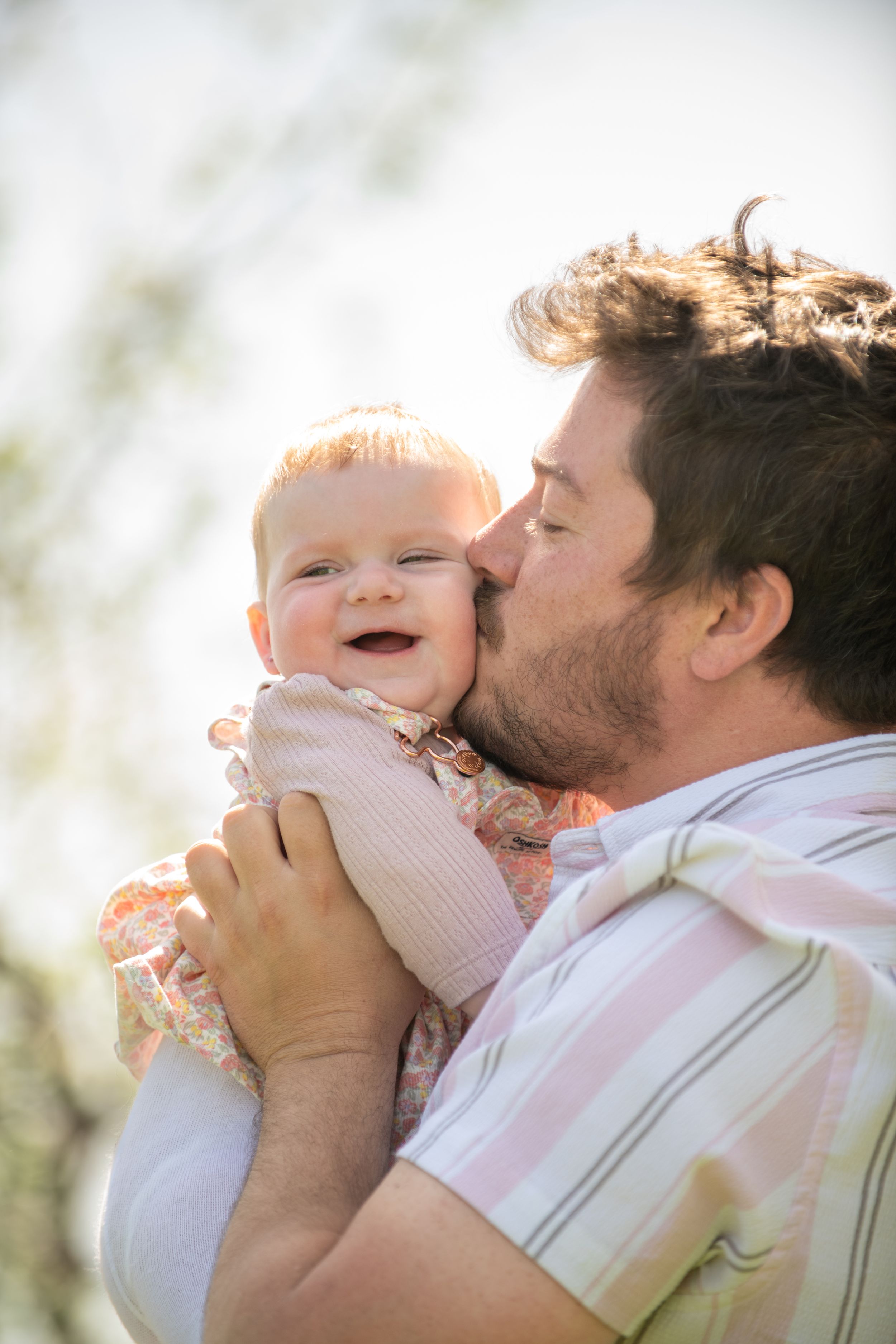 A loving father kisses his smiling baby girl outdoors in warm sunlight, holding her gently in his arms.