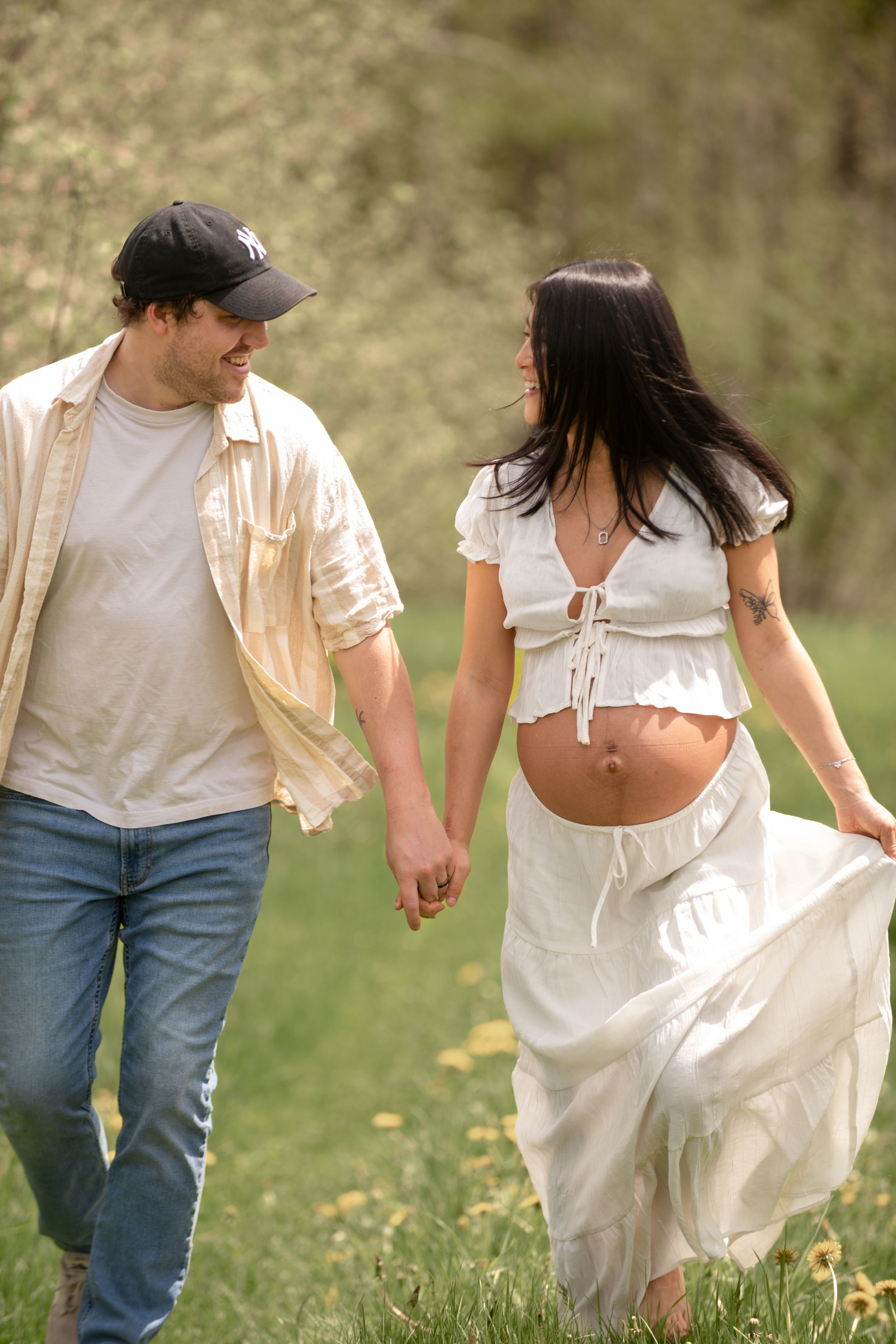 Pregnant woman in white crop top and skirt holds hands with man in cap and jeans walking outdoors.