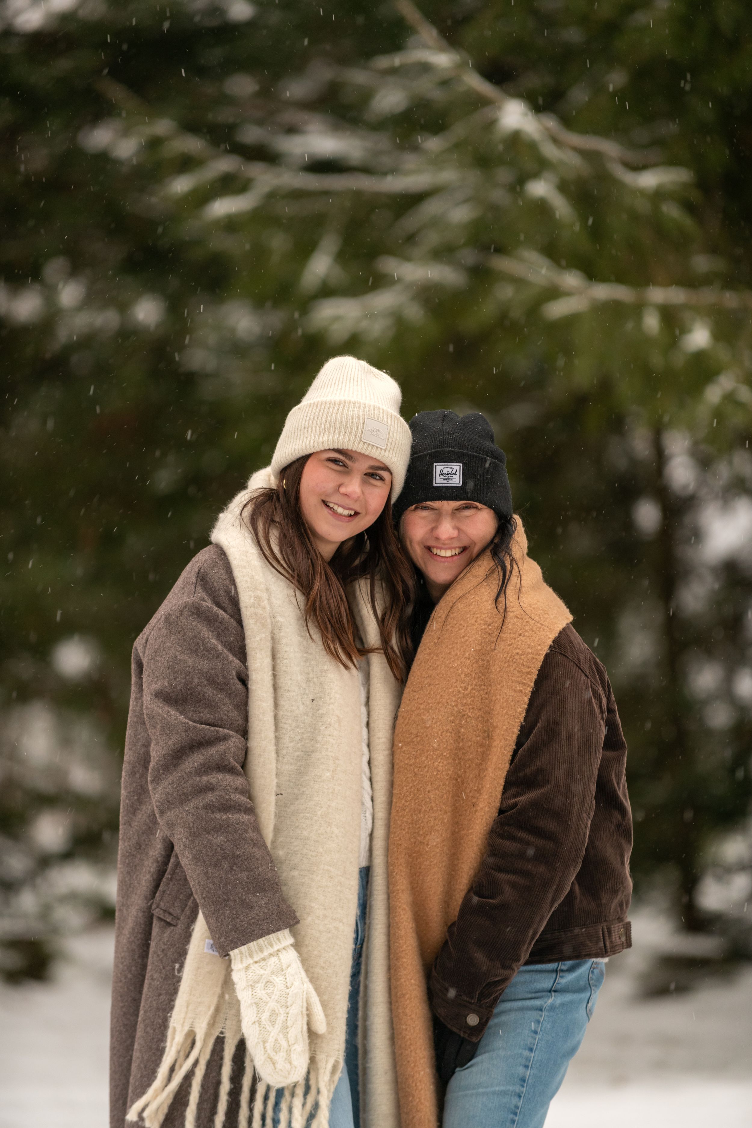Two friends bundled up in winter coats, beanies and scarves smile together in the falling snow.