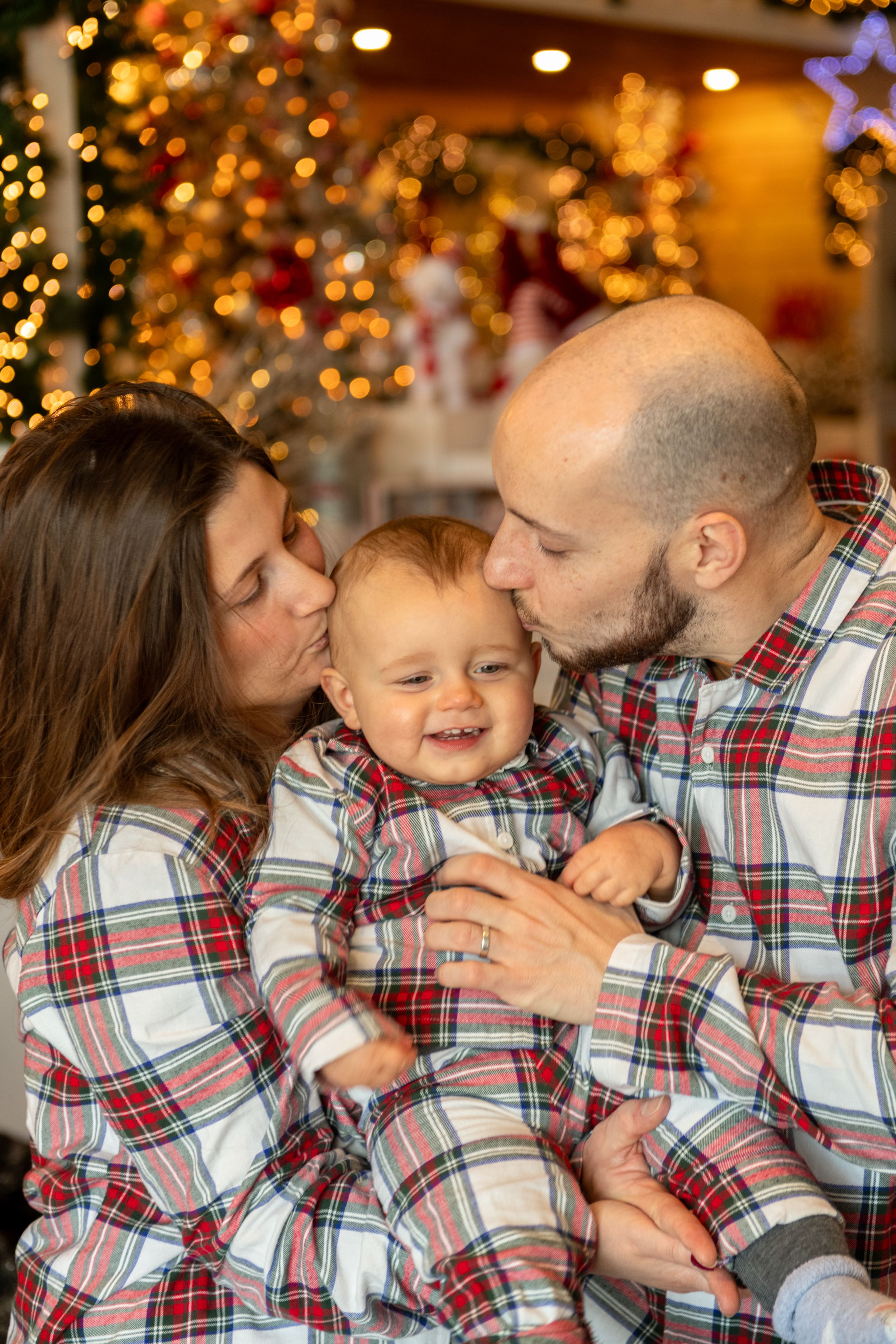 A family wearing matching plaid pajamas shares playful kisses and laughter in front of Christmas lights.