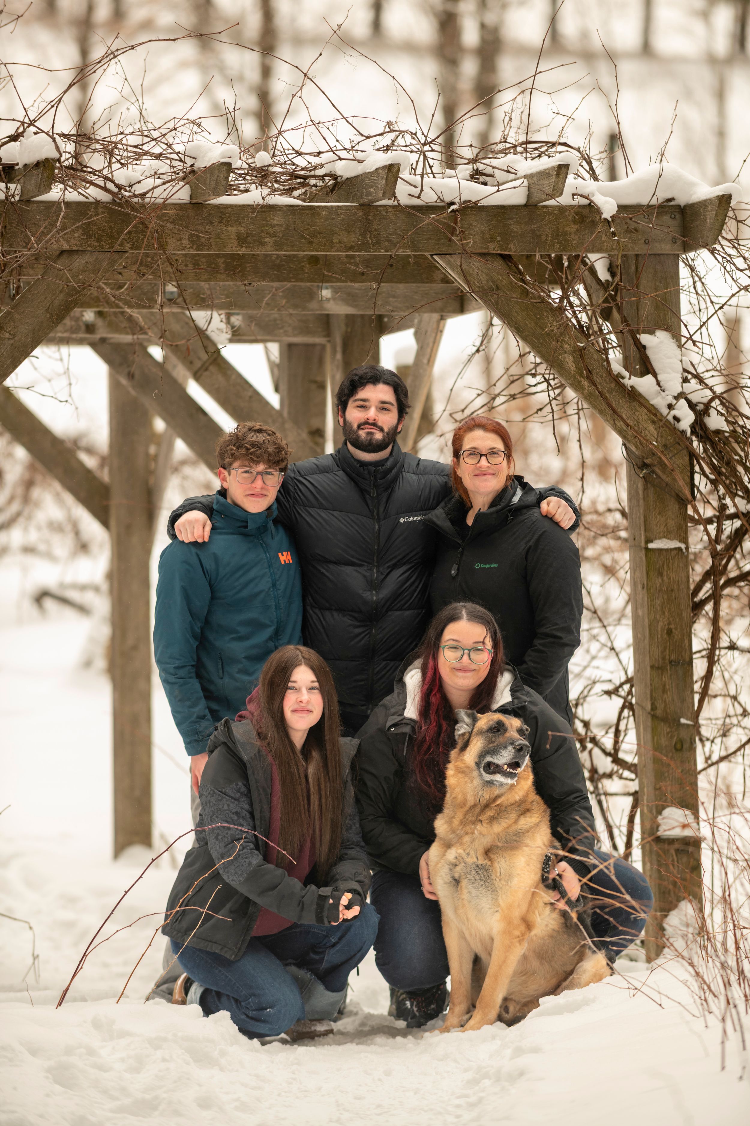 Family of five poses with German Shepherd dog under snowy wooden pergola in winter landscape.