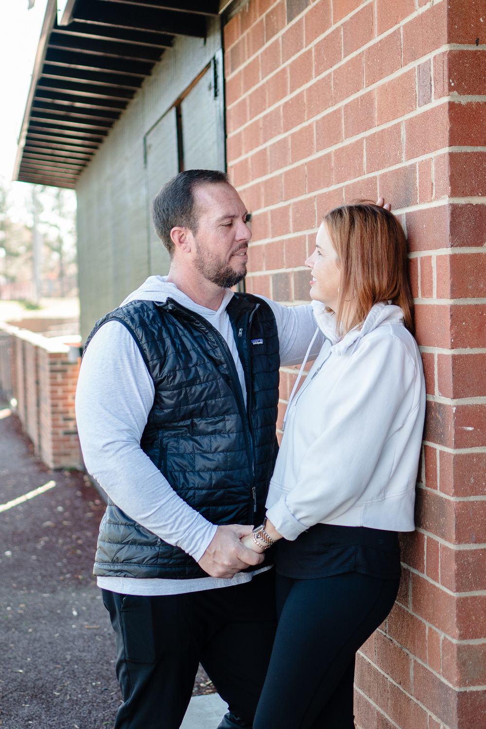 A couple shares an intimate moment against a brick wall exterior, with one wearing a black vest and the other in white.