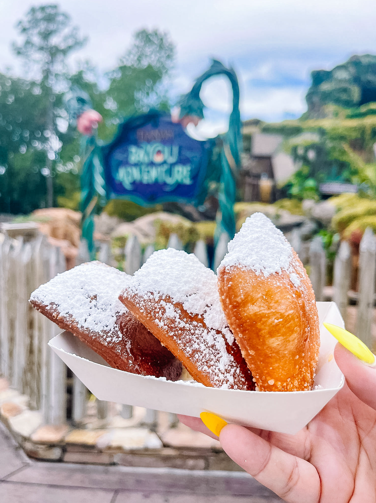 Deep-fried beignets dusted with powdered sugar held in front of a Jurassic-themed attraction sign at a theme park.