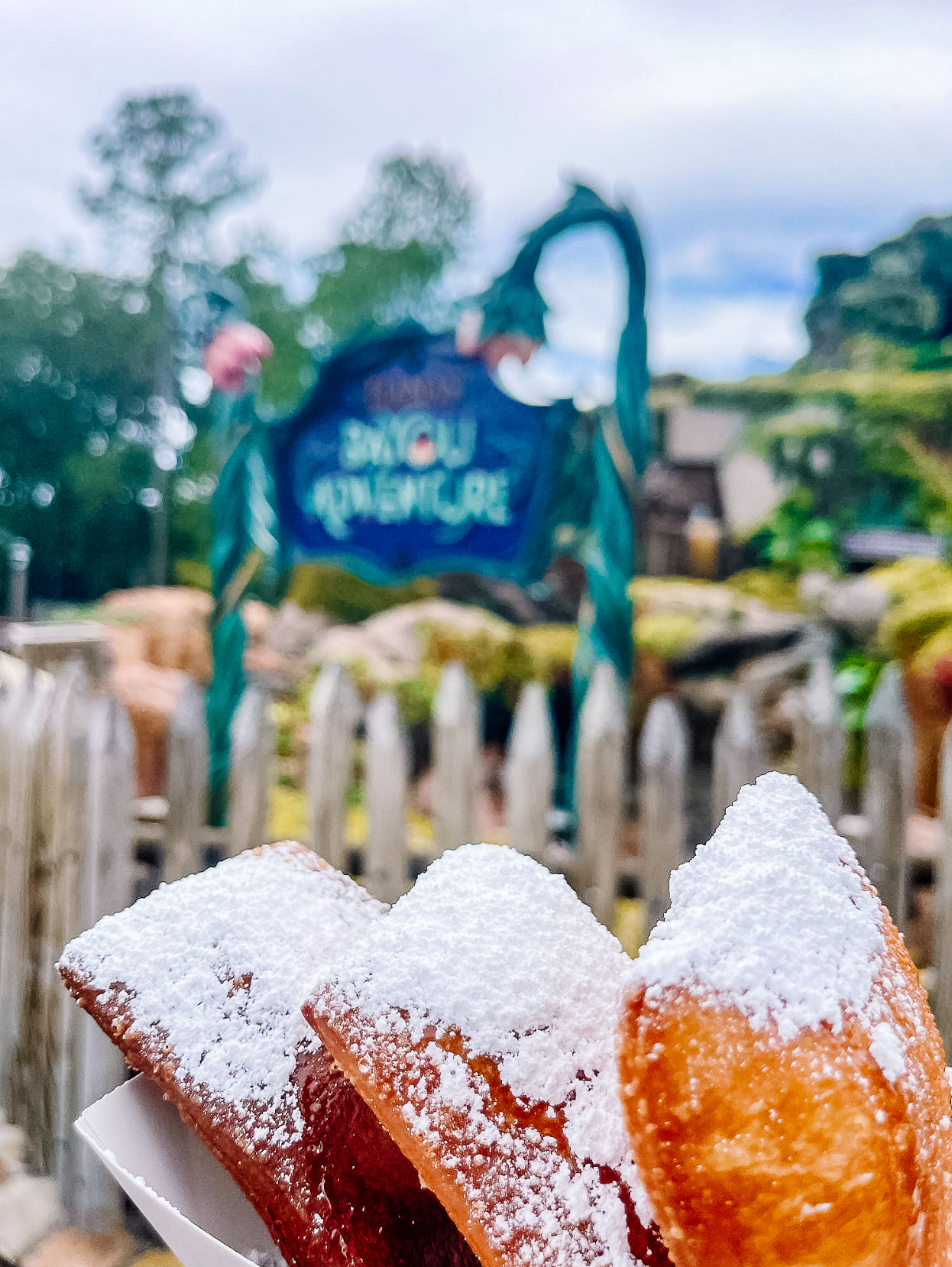 Powdered sugar is sprinkled on top of deep-fried beignets against a blurred outdoor background with blue flags.