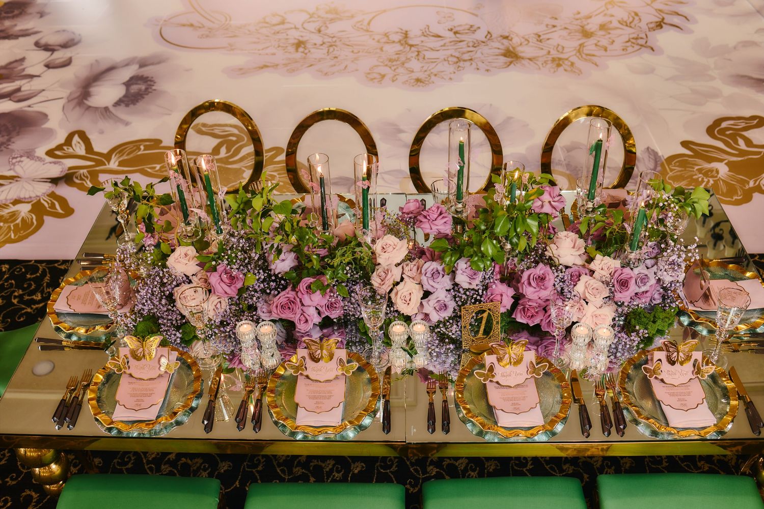 Elegant wedding table setting with pink floral centerpieces, gold chairs and crystal glassware on a green velvet table.