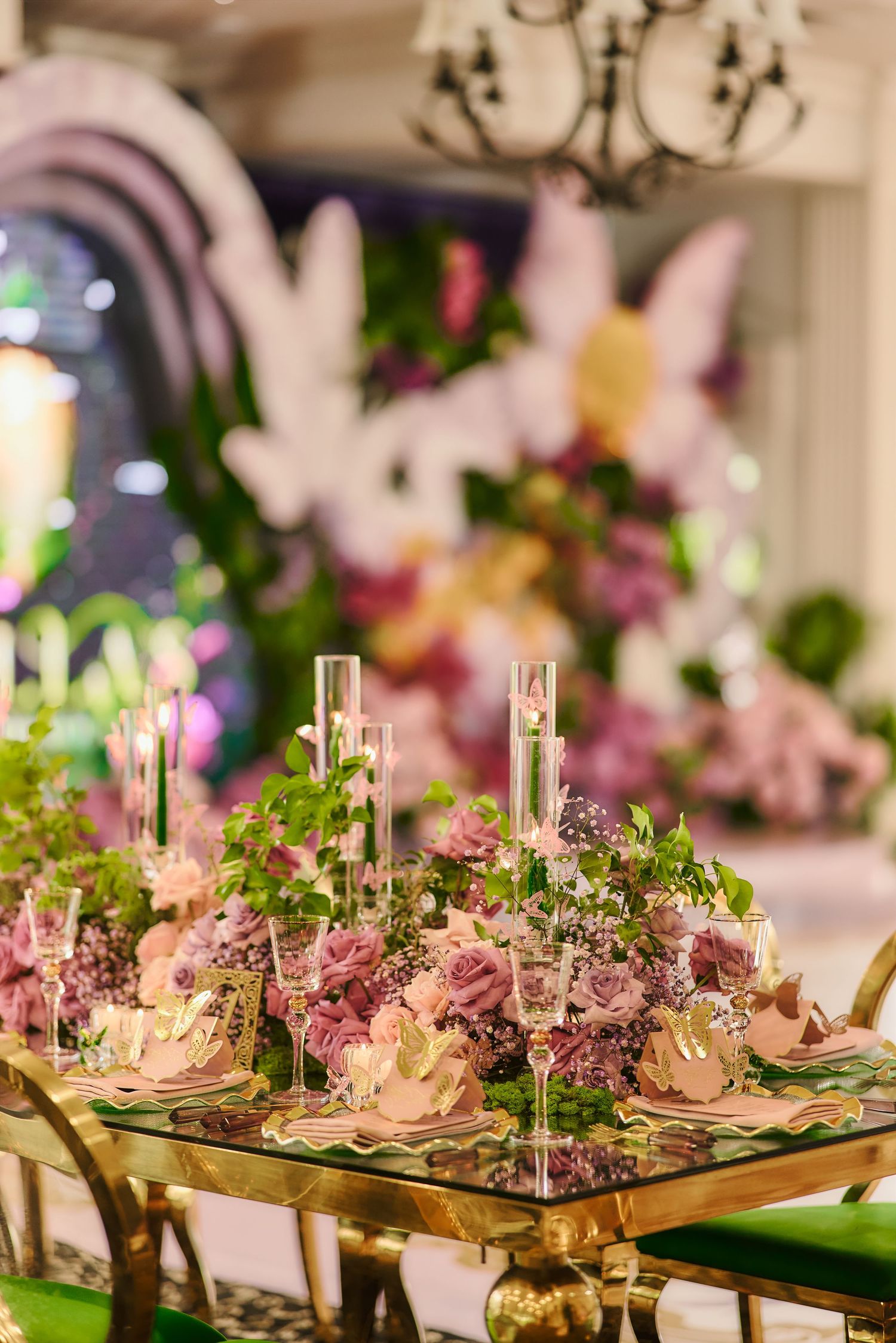 Elegant wedding table setting with pink floral centerpieces, tall glass candle holders and gold chairs in a luxurious venue.