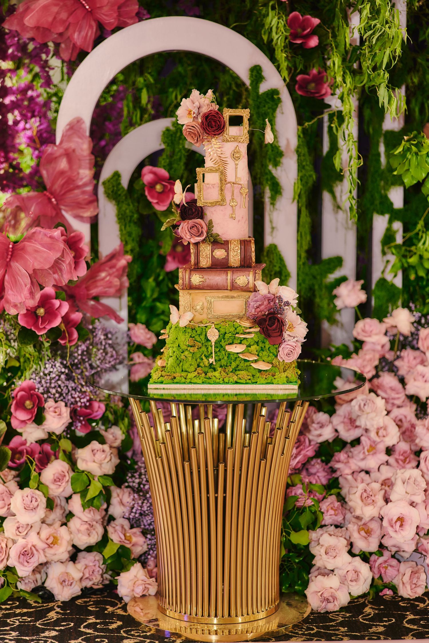 Elegant gold and pink wedding cake surrounded by cascading roses and greenery on black marbled surface.