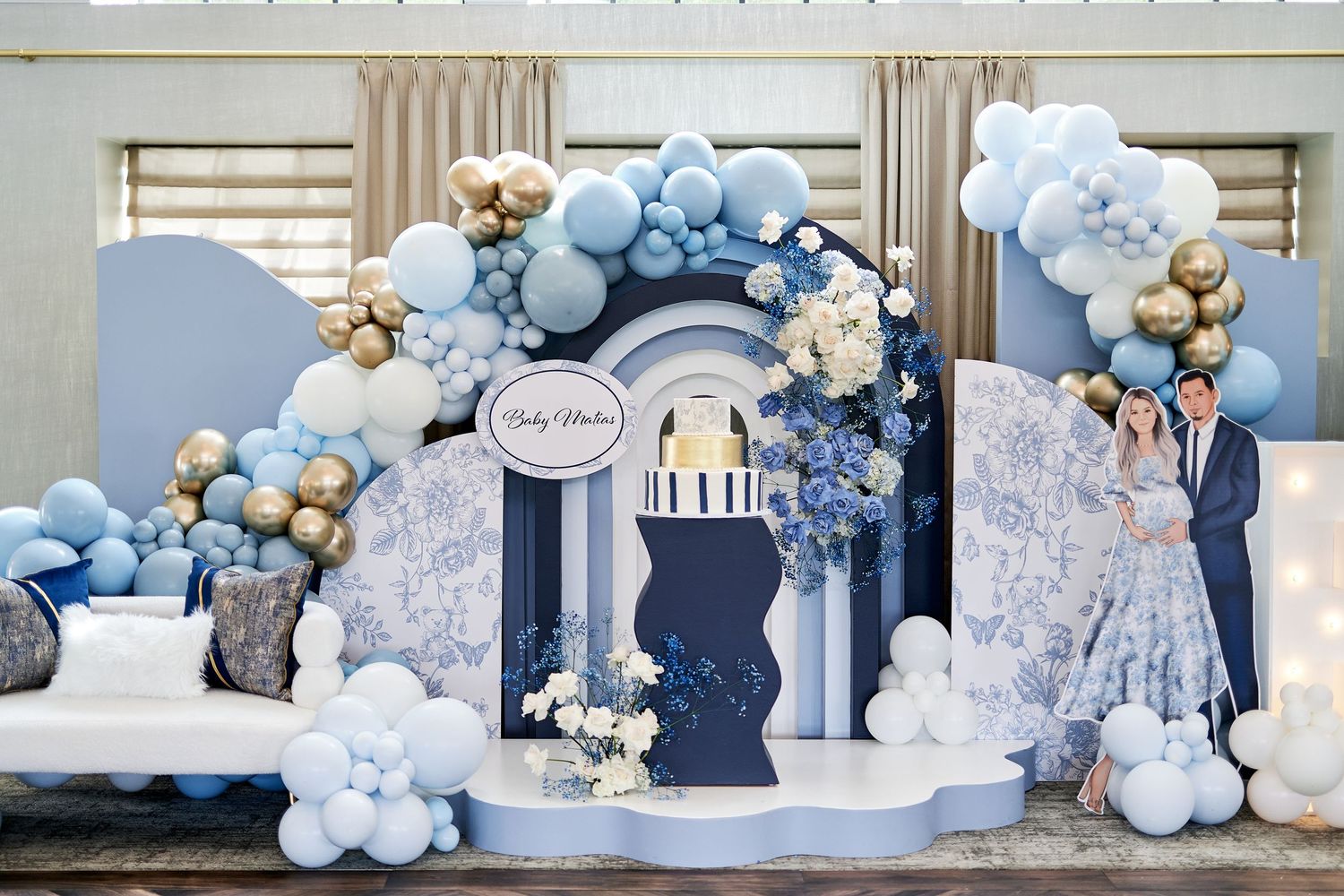 Elegant baby shower backdrop with blue and gold balloon arch, floral arrangements, and decorative arches in navy and white.