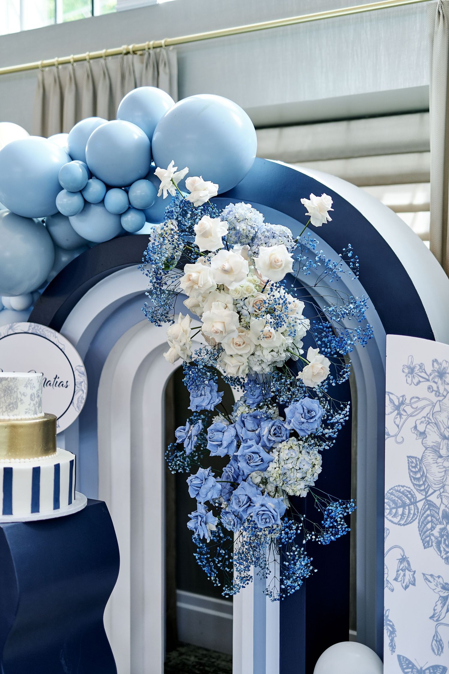 Elegant blue and white party decoration with balloon arch, floral arrangements and striped cake display against gray arches.