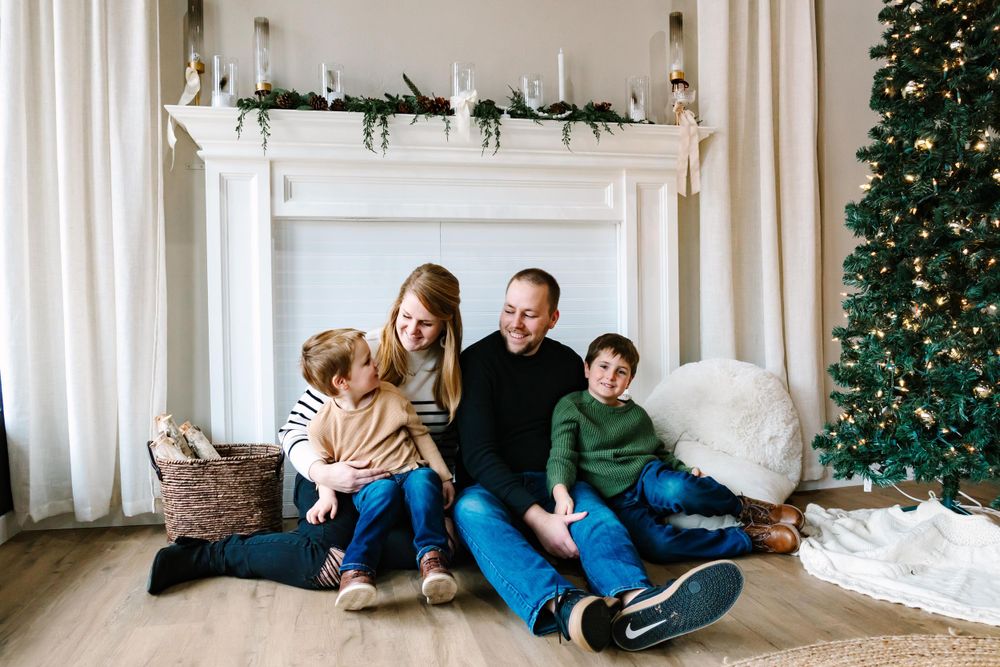 Family sits together in cozy living room near decorated Christmas tree and white fireplace mantel with garland.