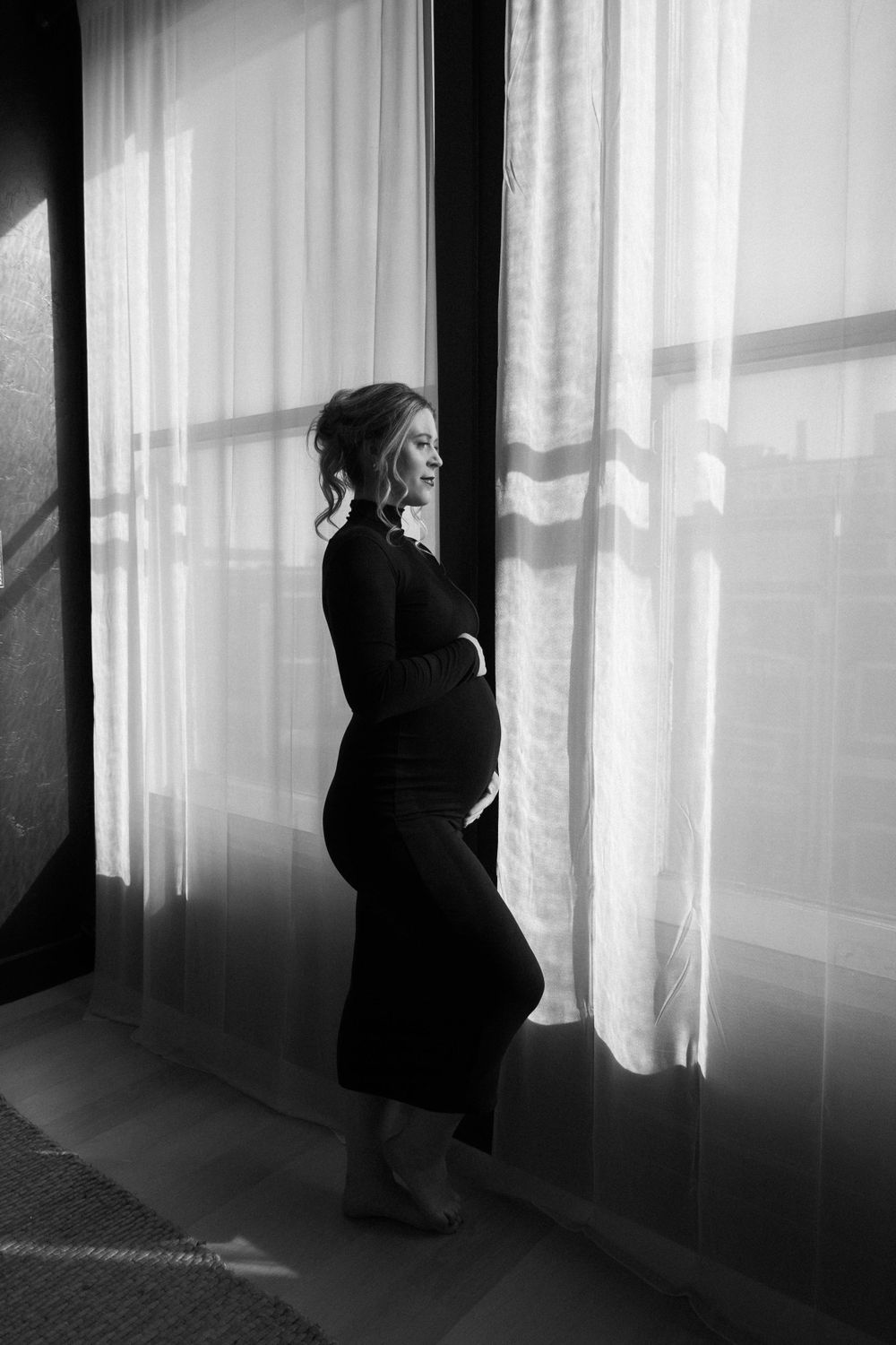 Artistic black and white silhouette of a pregnant woman standing by sheer curtained windows in contemplative pose.
