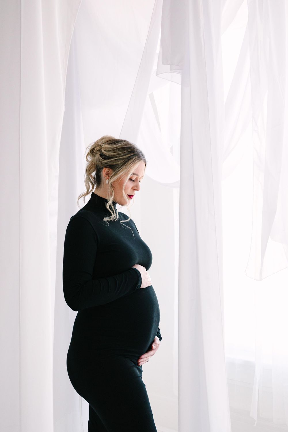 Silhouetted against sheer white curtains, an expectant mother in a black dress gently cradles her belly.