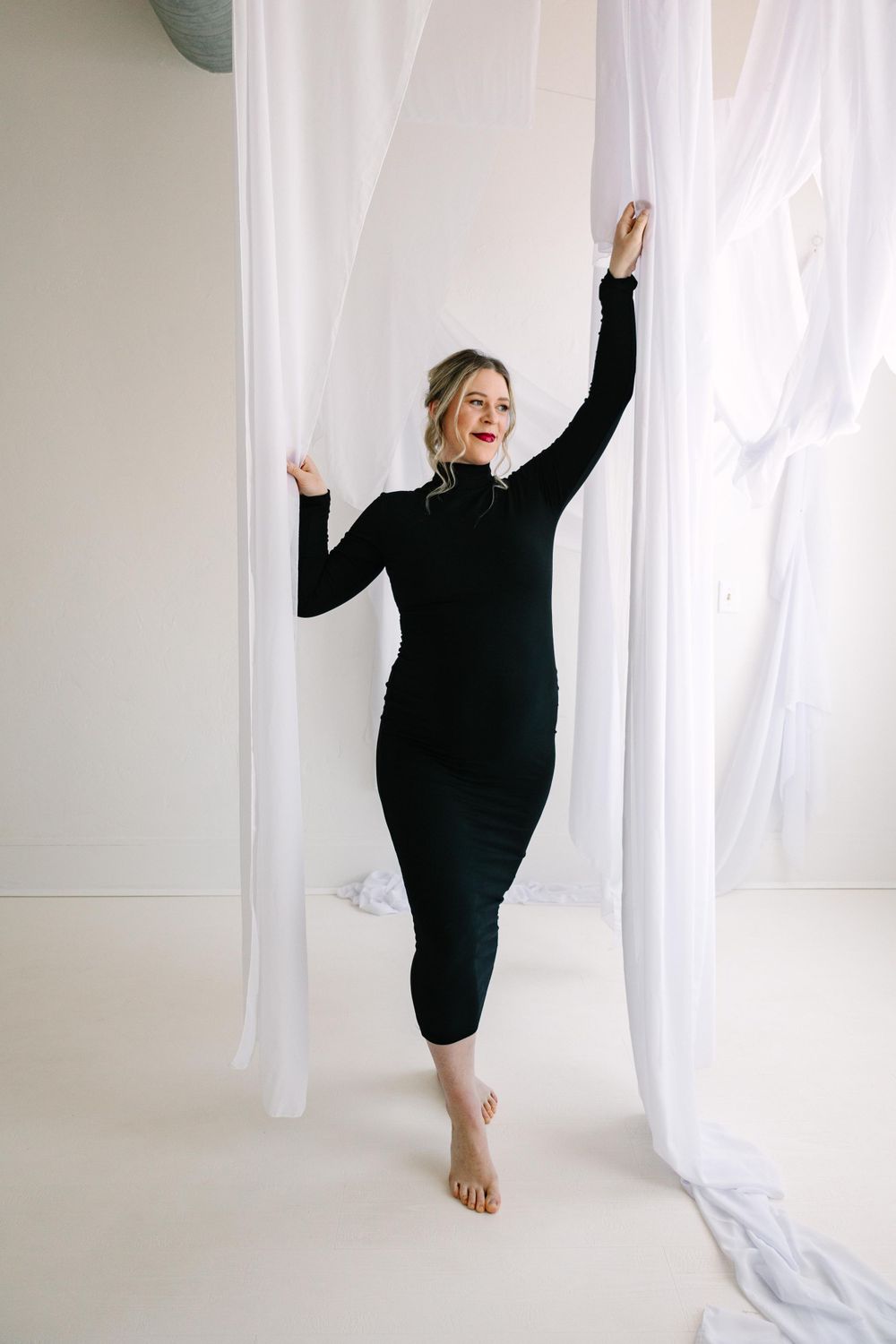 Woman in black long dress dancing gracefully between white curtains in a bright minimalist setting.