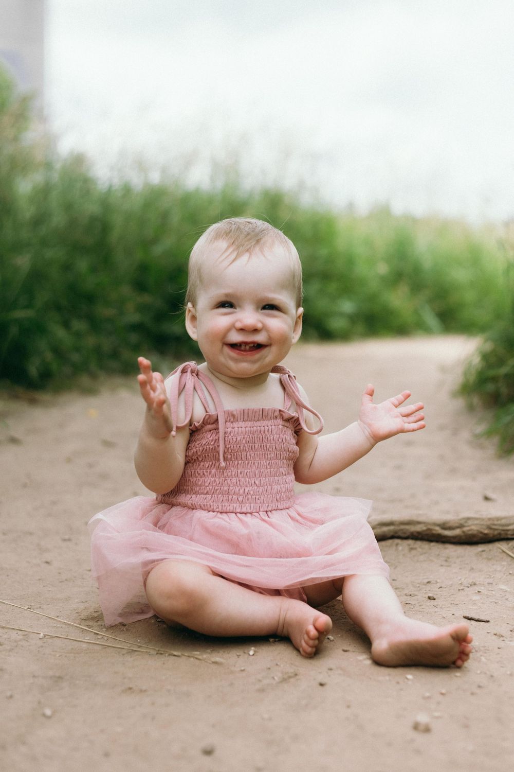 Baby in pink dress sitting on dirt path smiling with hands raised against nature background.