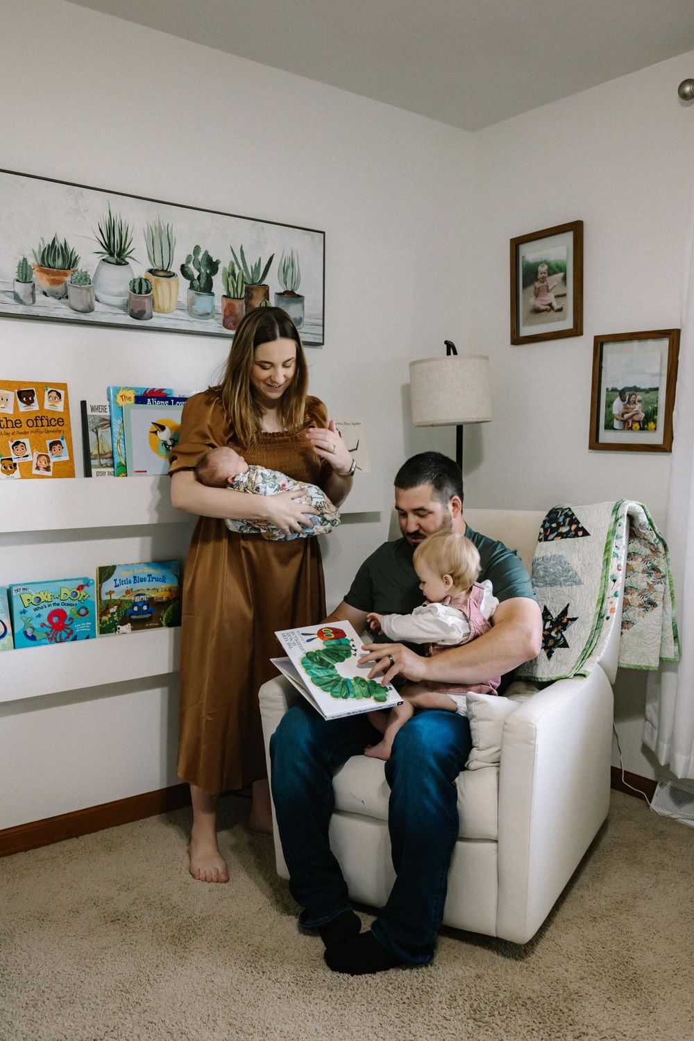 Family enjoys reading time together in cozy nursery corner with floating shelves, plants and artwork on white walls.