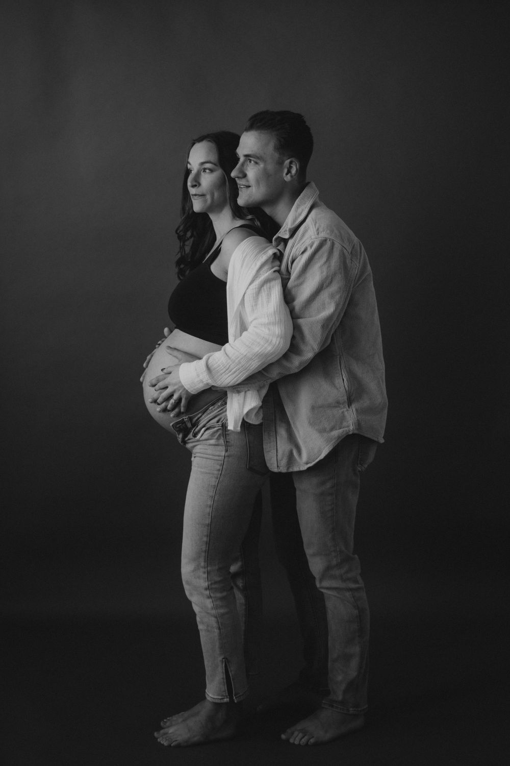 An expectant couple embraces in a tender black and white portrait against a dark background.