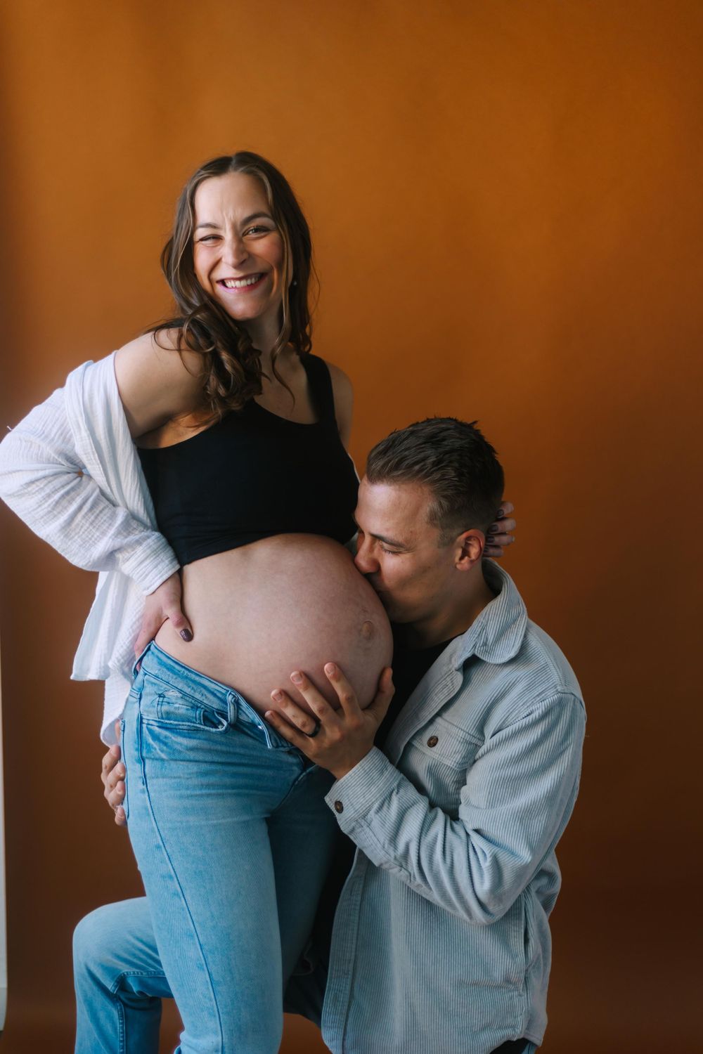 Maternity photo of expectant couple in casual attire against warm orange backdrop.