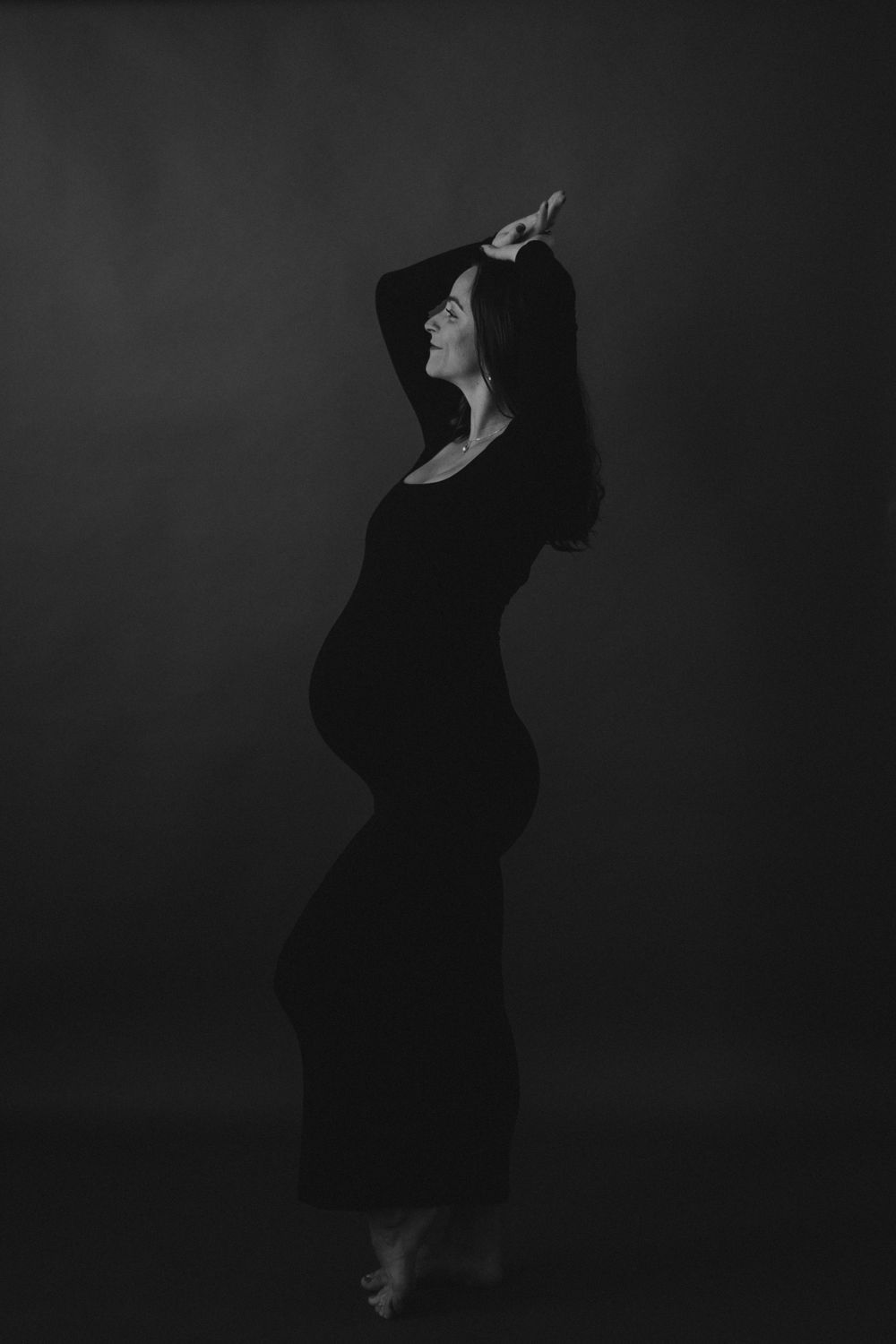 Artistic silhouette of an expectant mother in a black dress against dark background.
