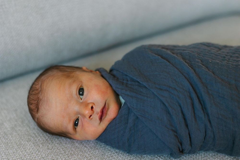 Baby swaddled in soft navy blue fabric laying on light gray surface while gazing at camera.