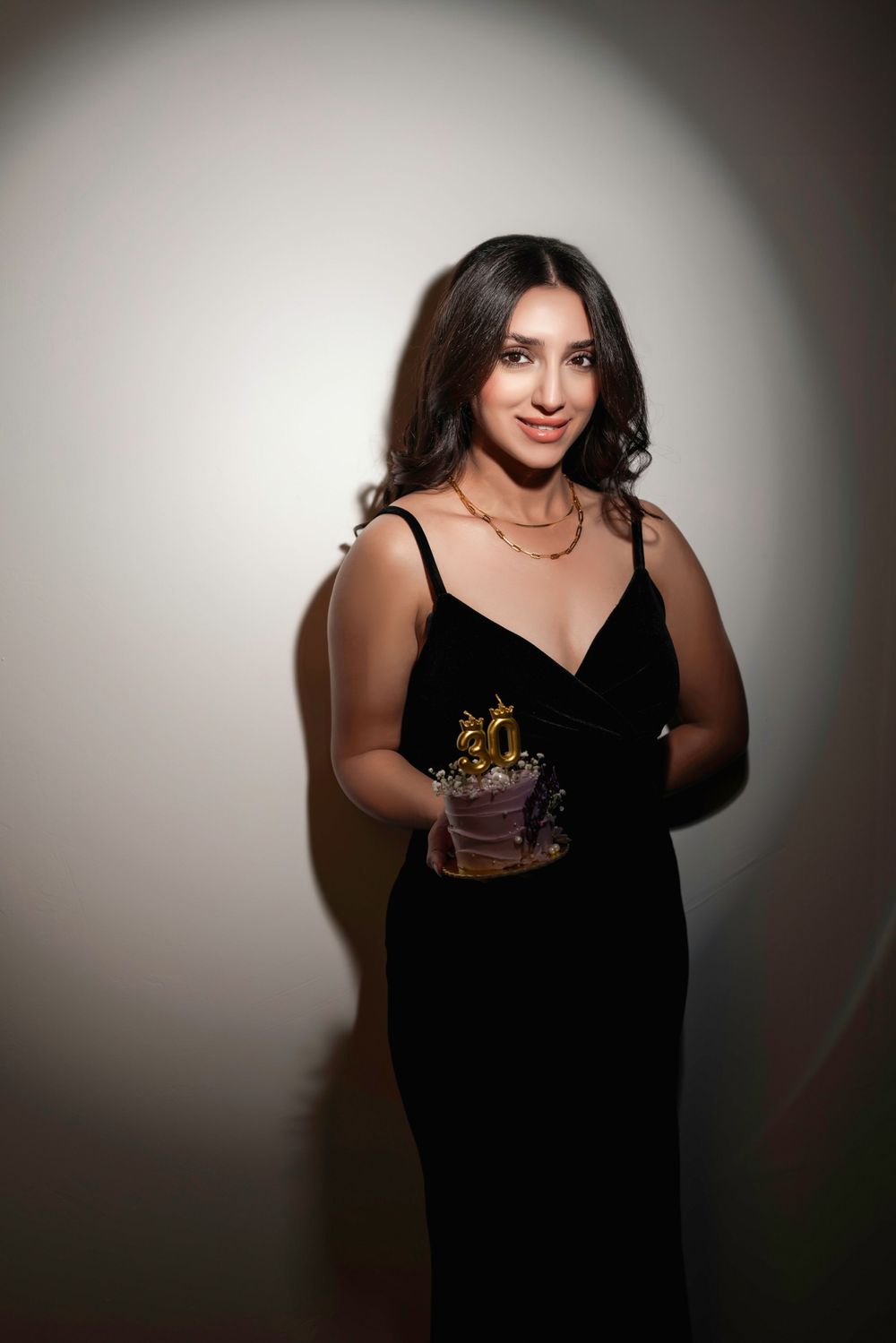 Woman in elegant black evening gown holds a small birthday cake in dramatic lighting against white background.