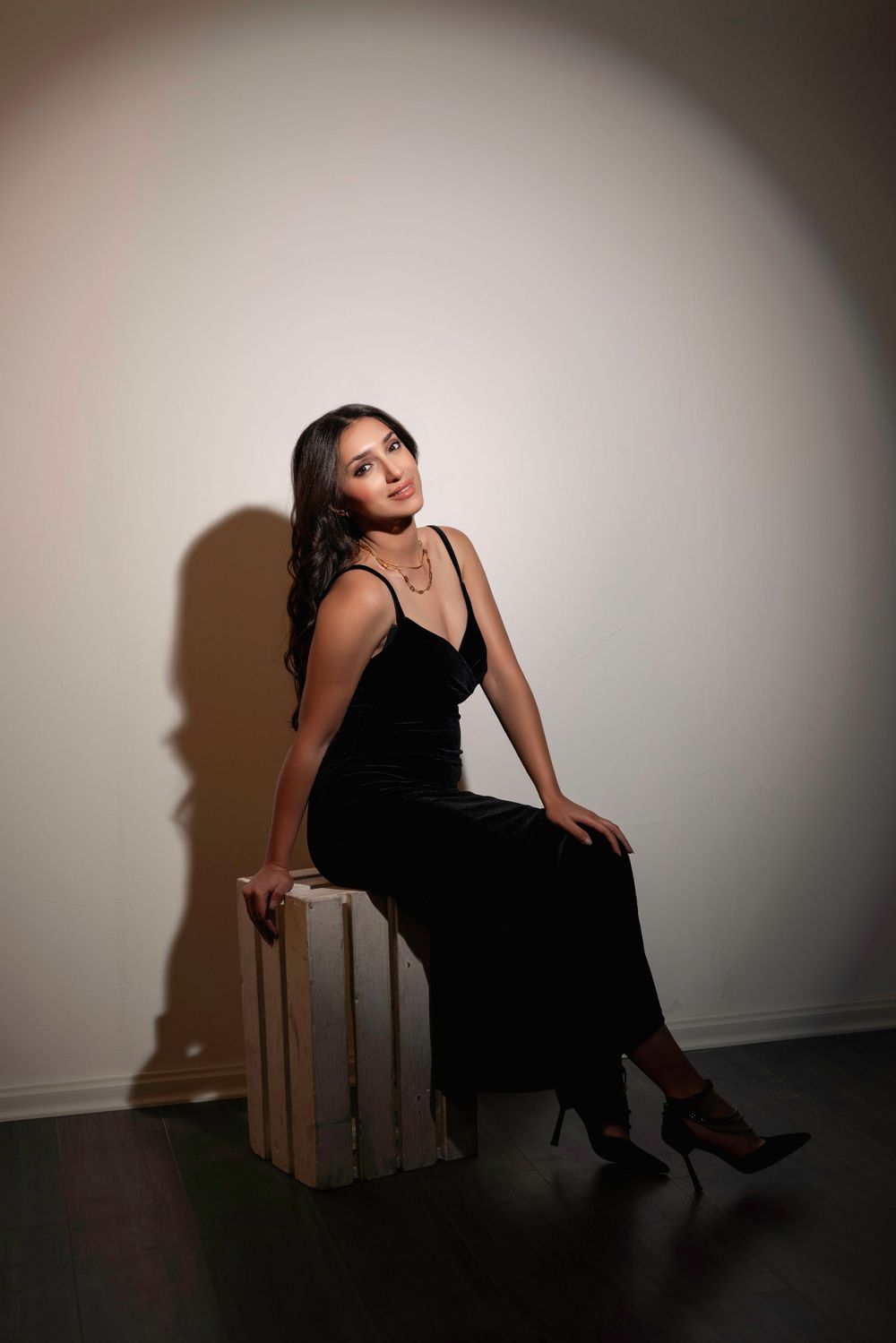 Woman in elegant black evening gown holds a small birthday cake in dramatic lighting against white background.