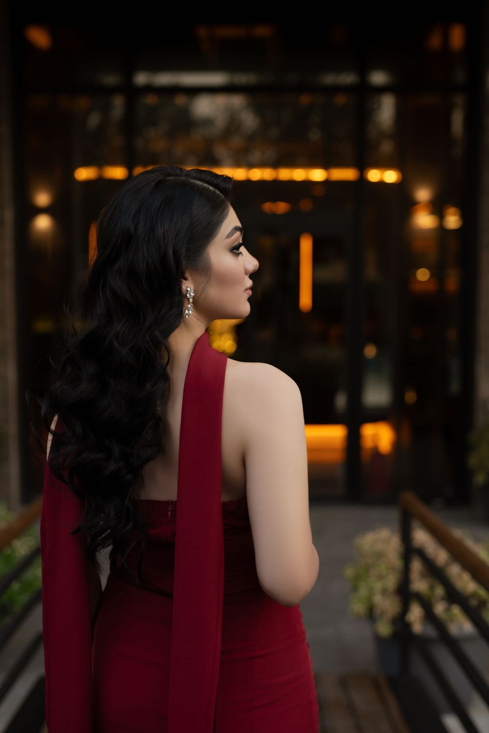 Woman in burgundy dress with dark hair poses elegantly against evening city backdrop with warm lighting.