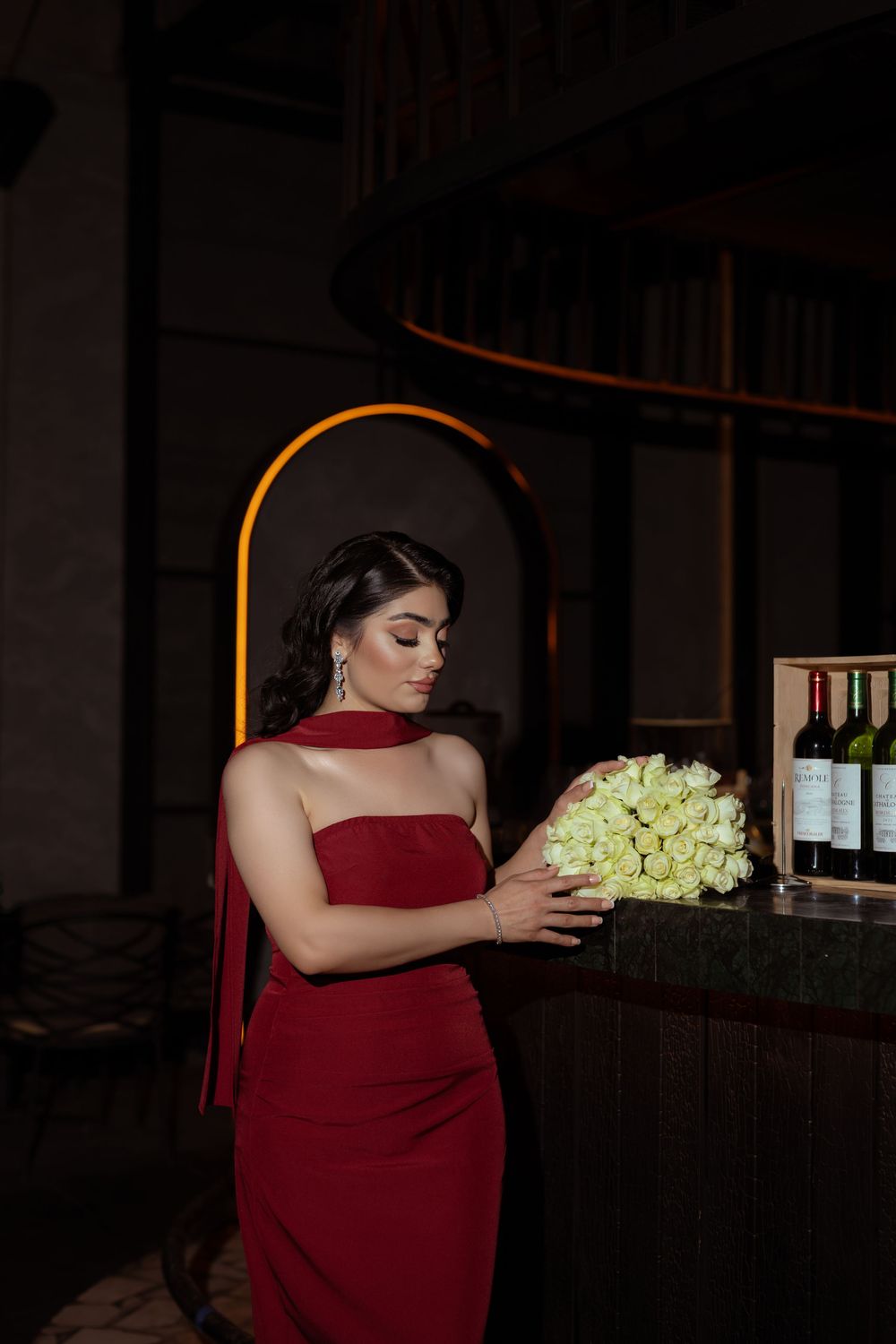 A person in a burgundy dress stands in a dimly lit room holding a bouquet of light-colored roses.