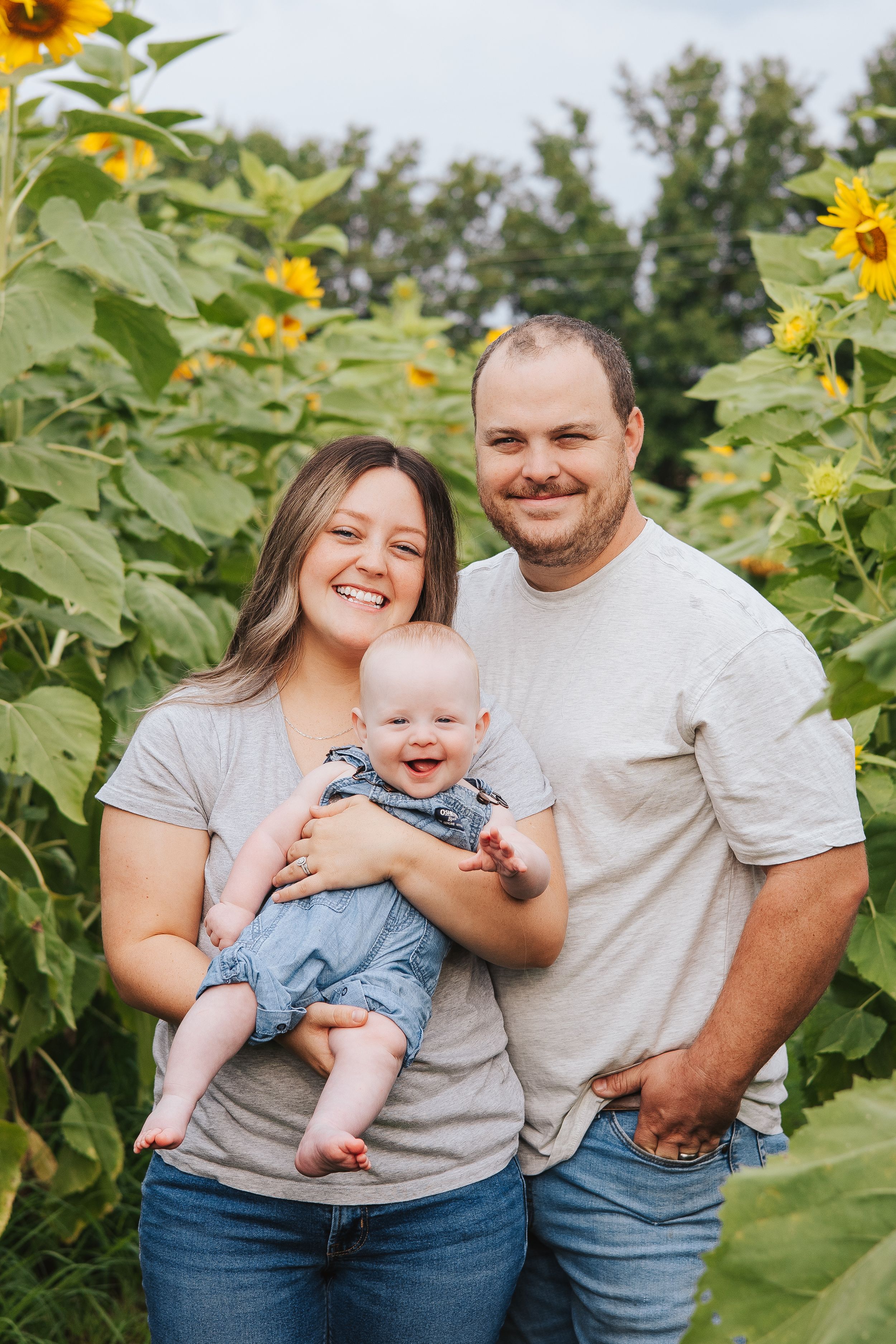 Baby in denim overalls smiles while being held in a sunflower field.