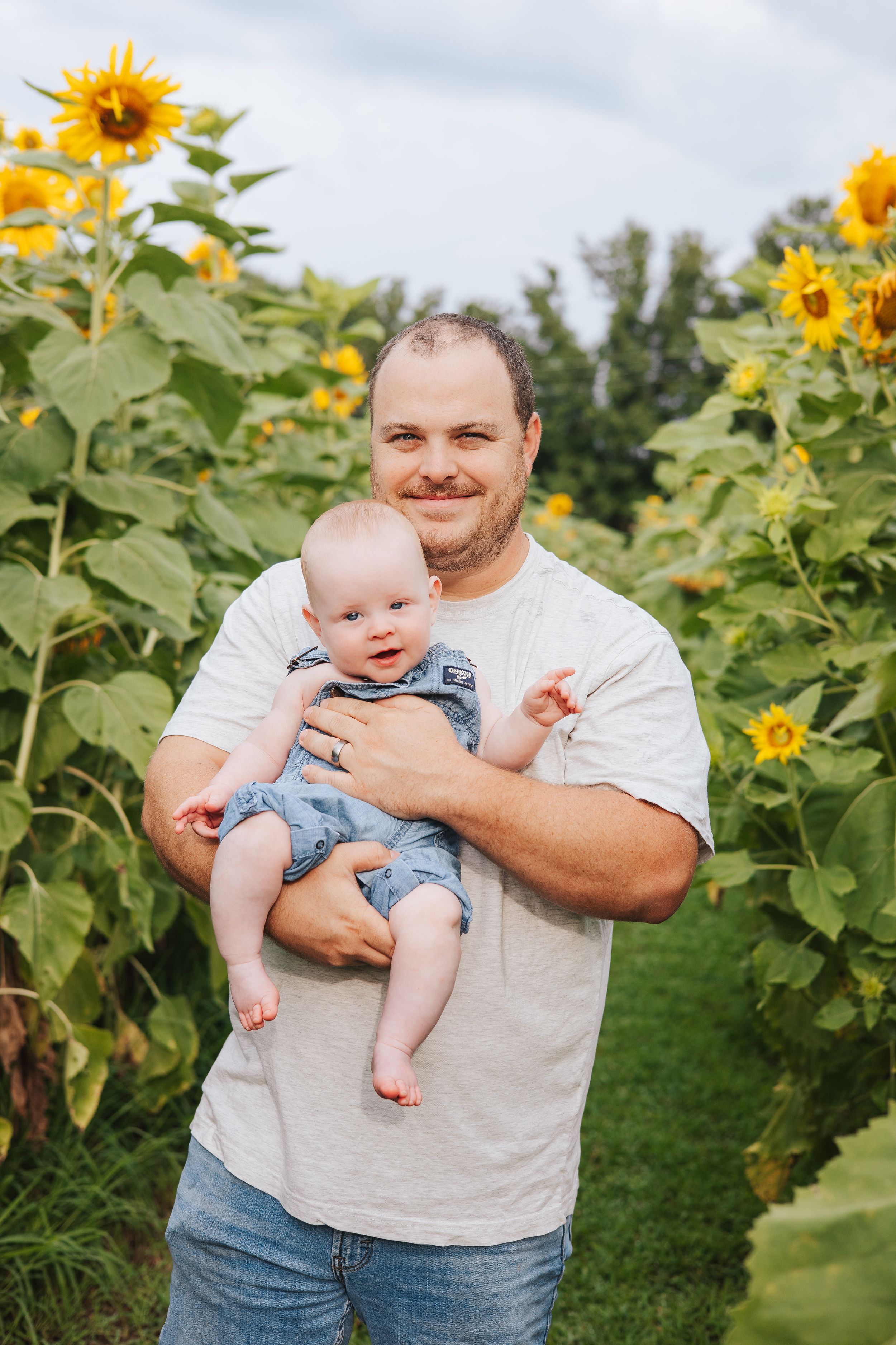 Baby in denim outfit held up against backdrop of towering sunflowers.