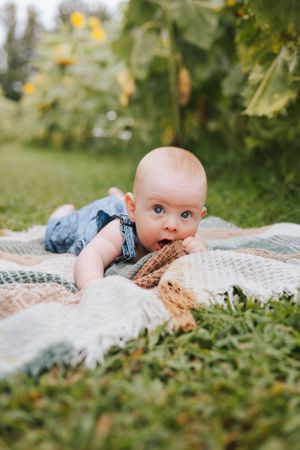 Baby in overalls lying on blanket outdoors exploring with mouth among sunflower garden.