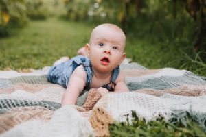 Baby in blue overalls crawling on blanket with curious expression in garden setting.