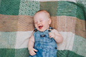 Joyful baby wearing denim overalls lying on geometric patterned blanket outdoors.