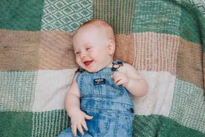 Baby in denim overalls laughing while lying on patchwork quilt with green and tan patterns.