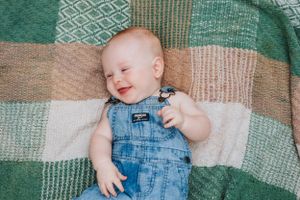 Baby in overalls laughing with pure joy while resting on patterned quilt.