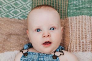 Baby in denim overalls with bright eyes looking up while lying on patterned blanket.