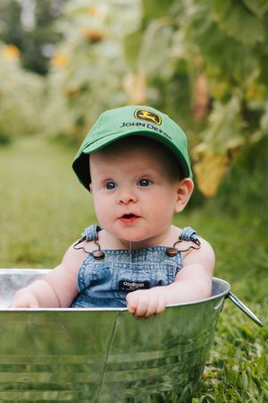 A small baby wearing overalls and a green cap sits in a metal wash tub outdoors.