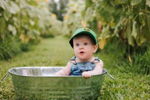 A baby in denim overalls and green hat peeks over the edge of a metal tub.