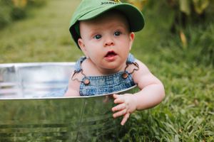 A baby in overalls and green cap explores a metal tub in an outdoor setting.