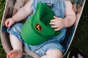 Small child holding a green John Deere baseball cap while wearing denim overalls.