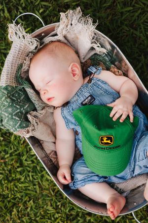 Sleeping baby in denim overalls holding a green John Deere cap in a basket outdoors.