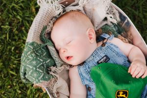 Peaceful sleeping baby in denim with John Deere cap resting in woven basket on grass.