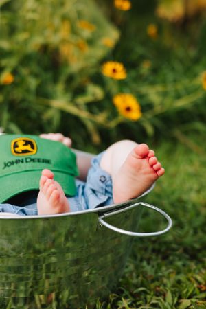 Tiny feet in a basket next to a green John Deere cap with yellow flowers in background.