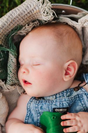 Sleeping baby in denim overalls with green cap resting in textured basket.