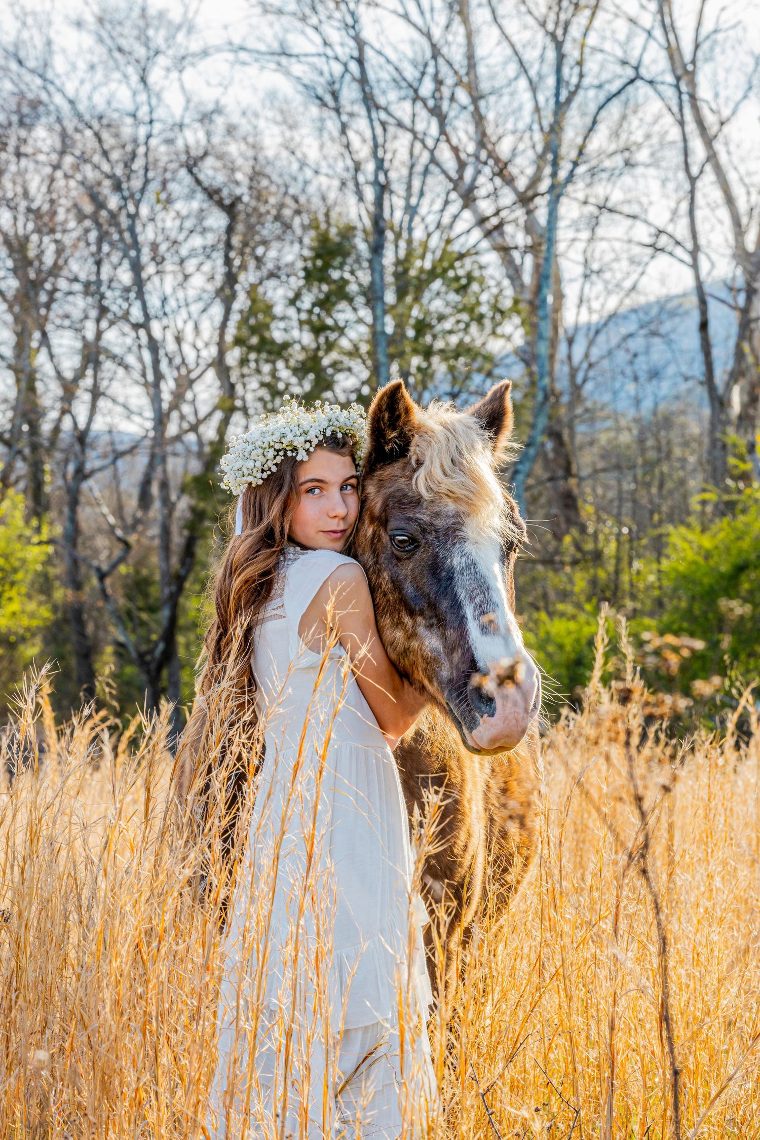Brown horse and white-dressed figure with flower crown pose together in golden autumn field at sunset.
