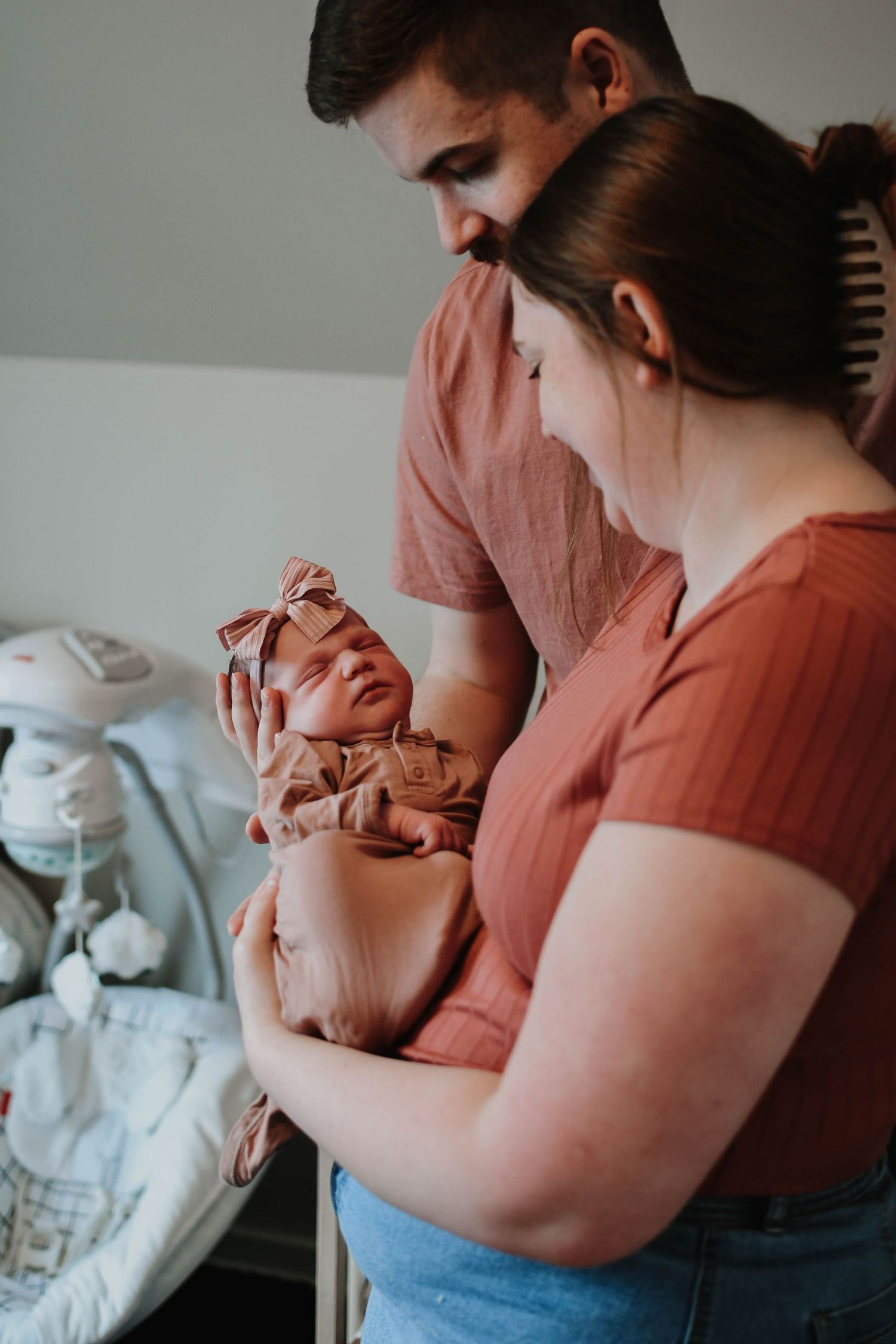 New parents in casual clothing tenderly hold their swaddled newborn baby in a hospital room setting.